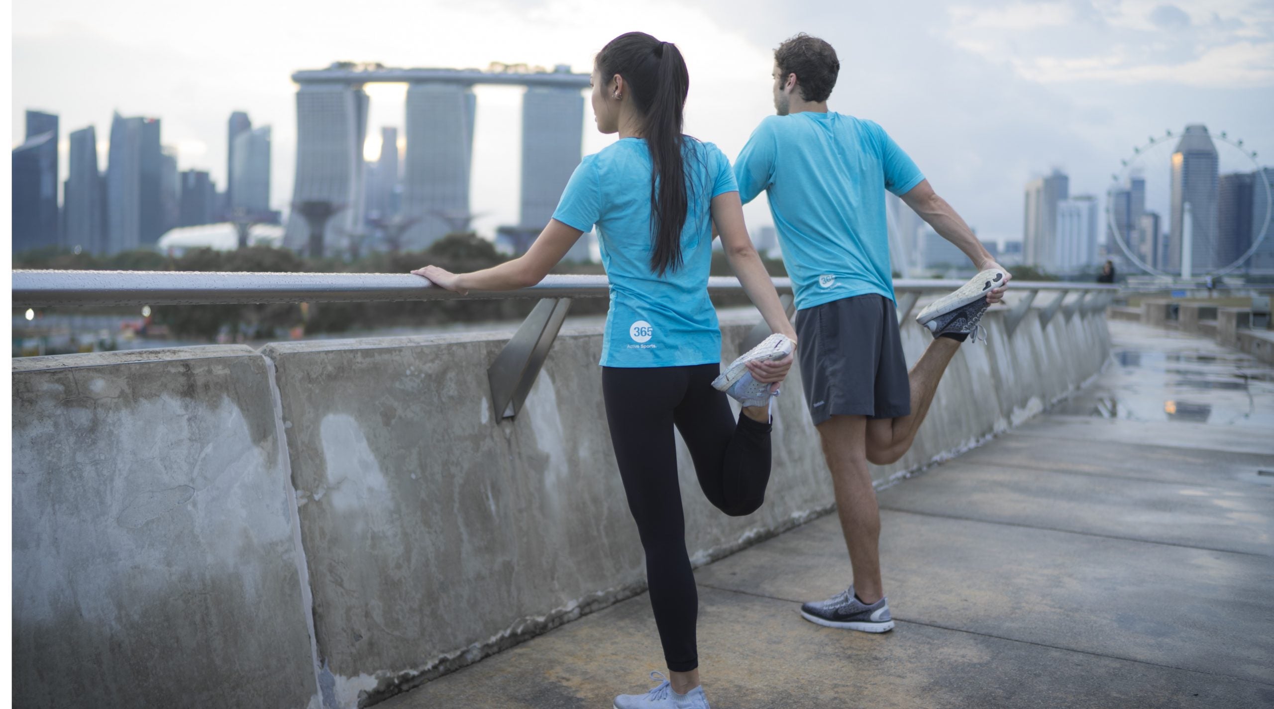 Two sports persons; one female and one male wearing blue t-shirts doing exercise while standing in front of towers building.