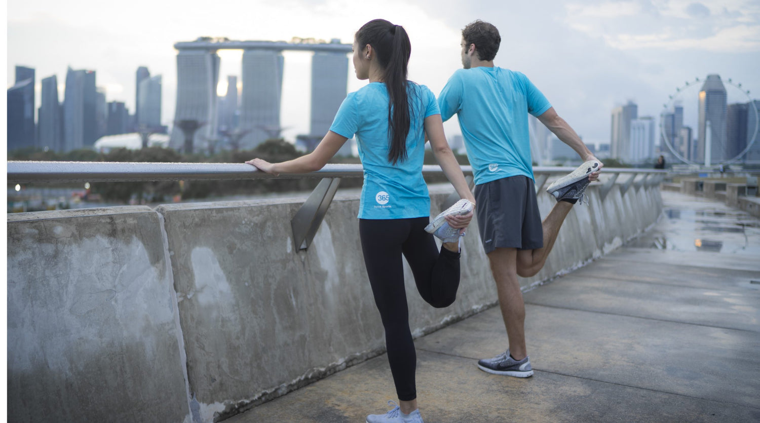 Two sports persons; one female and one male wearing blue t-shirts doing exercise while standing in front of towers building.